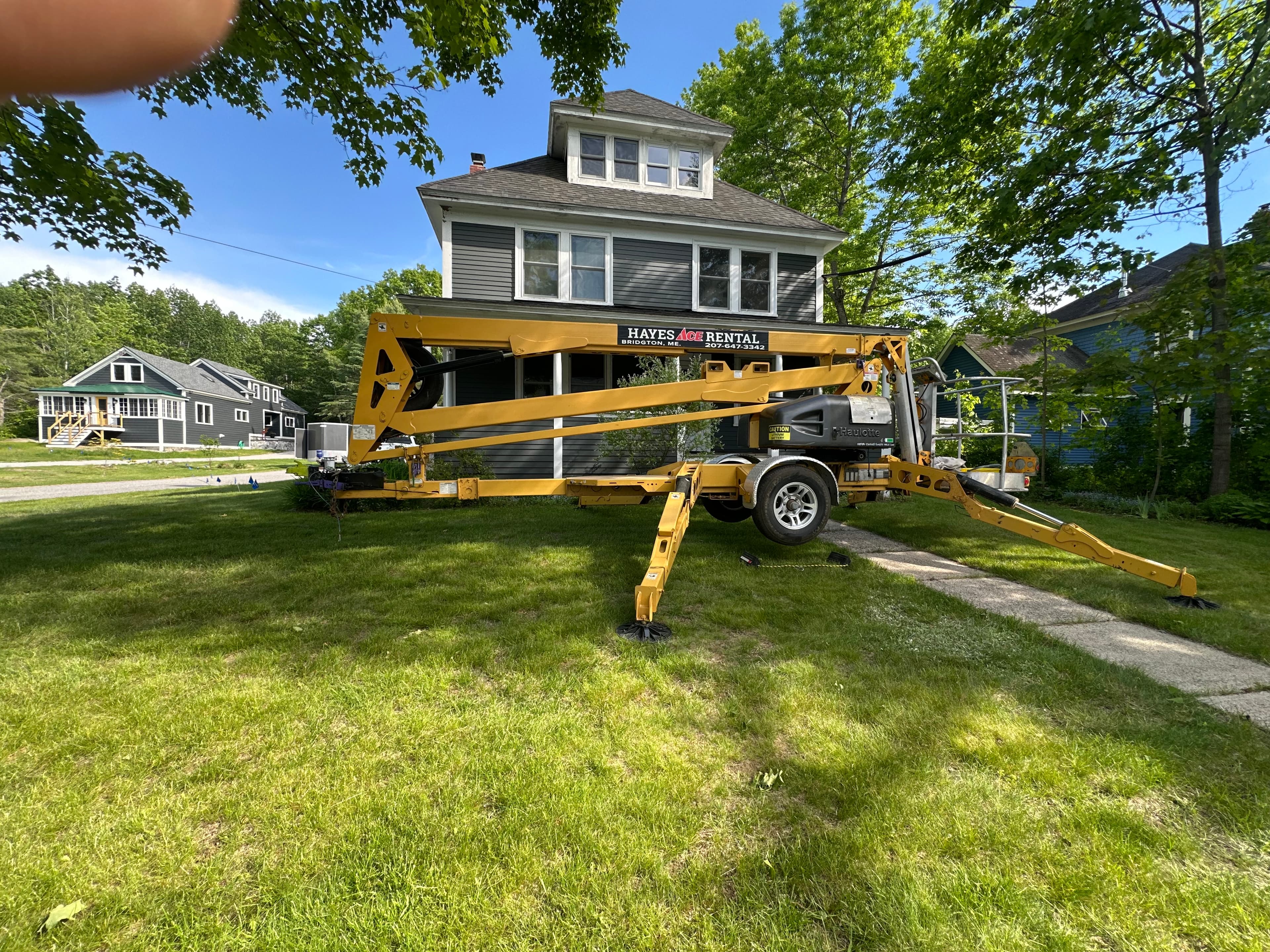 Yellow towable boom lift with outriggers extended on a lawn before a gray house.