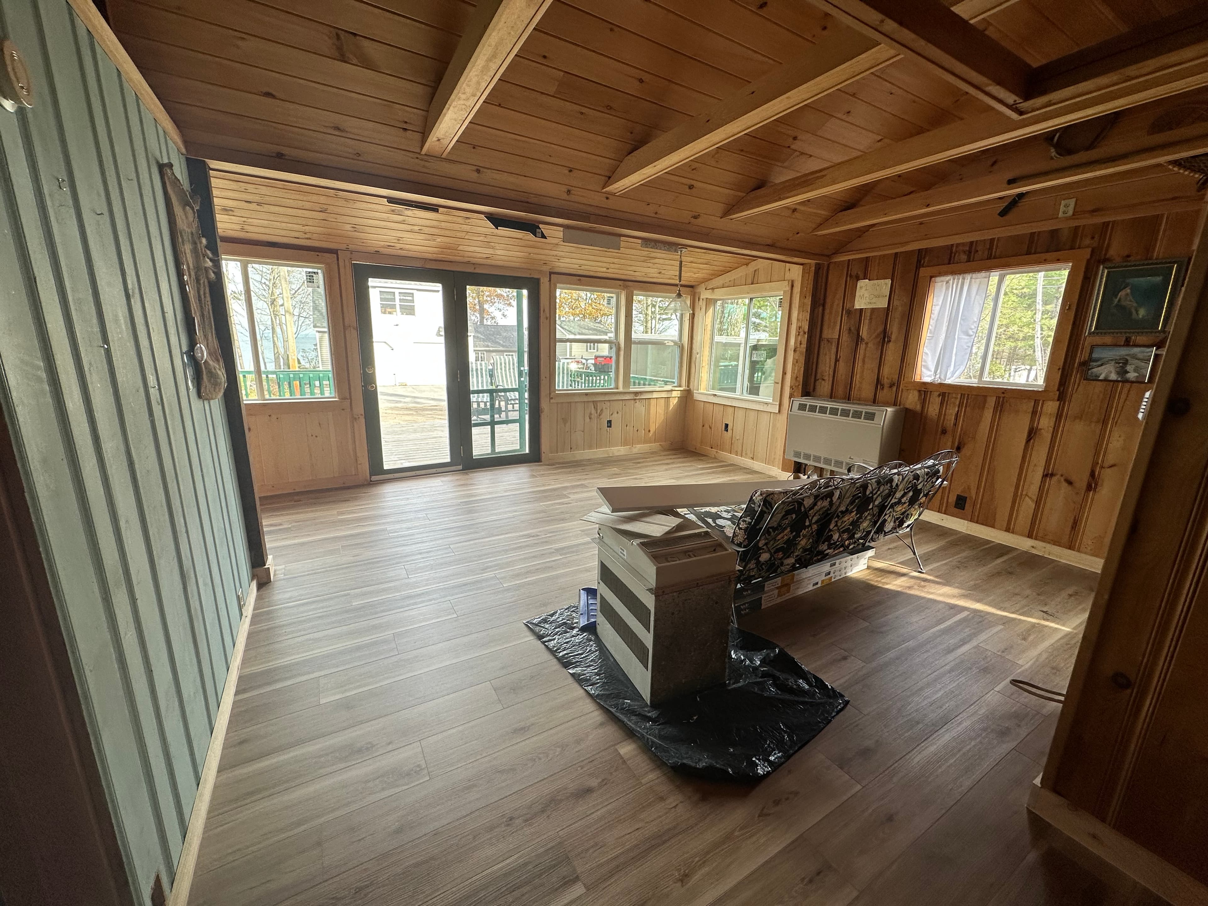 Rustic wood-paneled cabin interior with large windows, glass doors, and a sofa under renovation.
