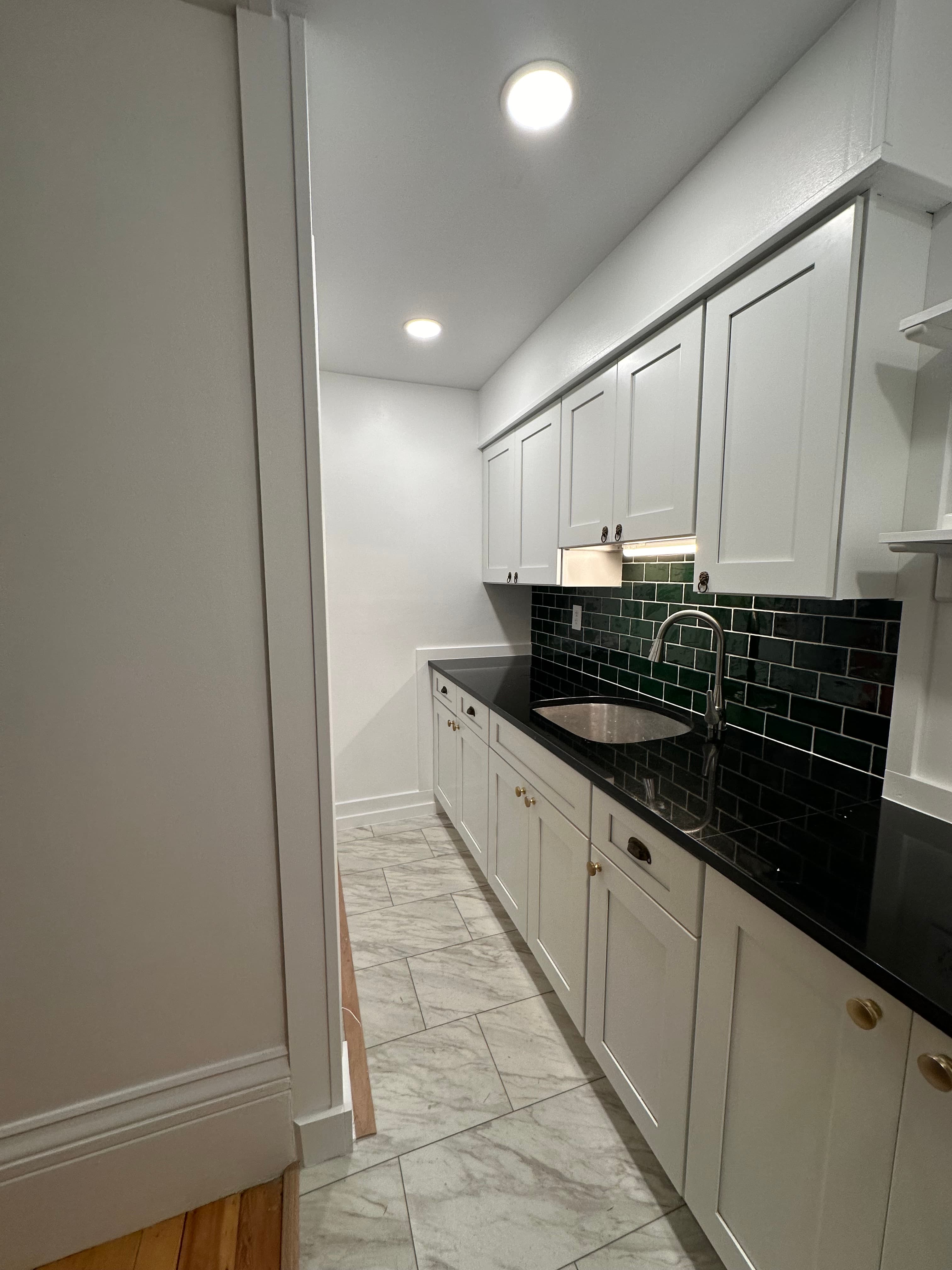 Narrow kitchen featuring white cabinets, black countertops, green subway tile backsplash, and marble floors.