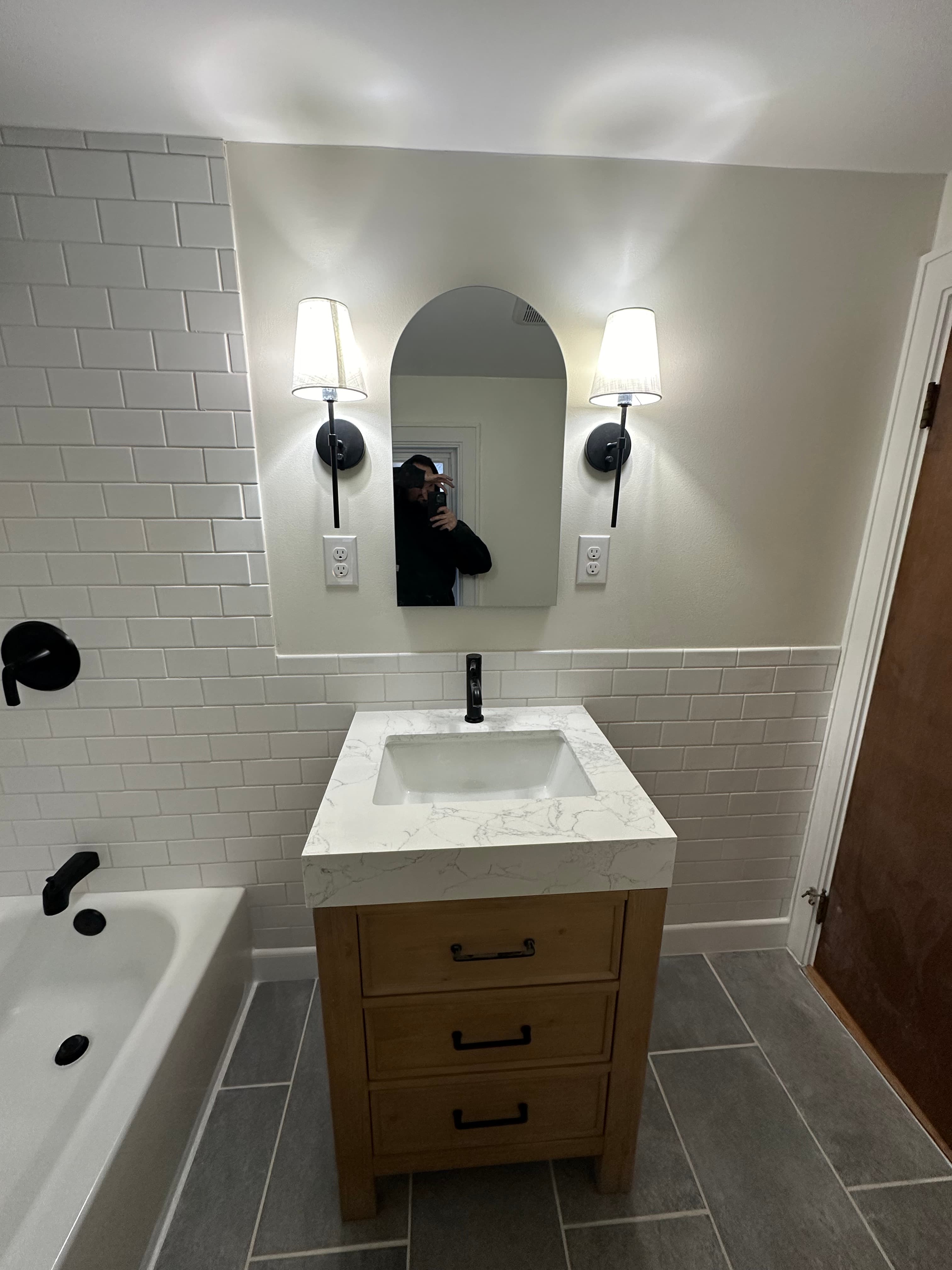 Modern wood vanity with marble top, arched mirror, and black fixtures in a tiled bathroom.