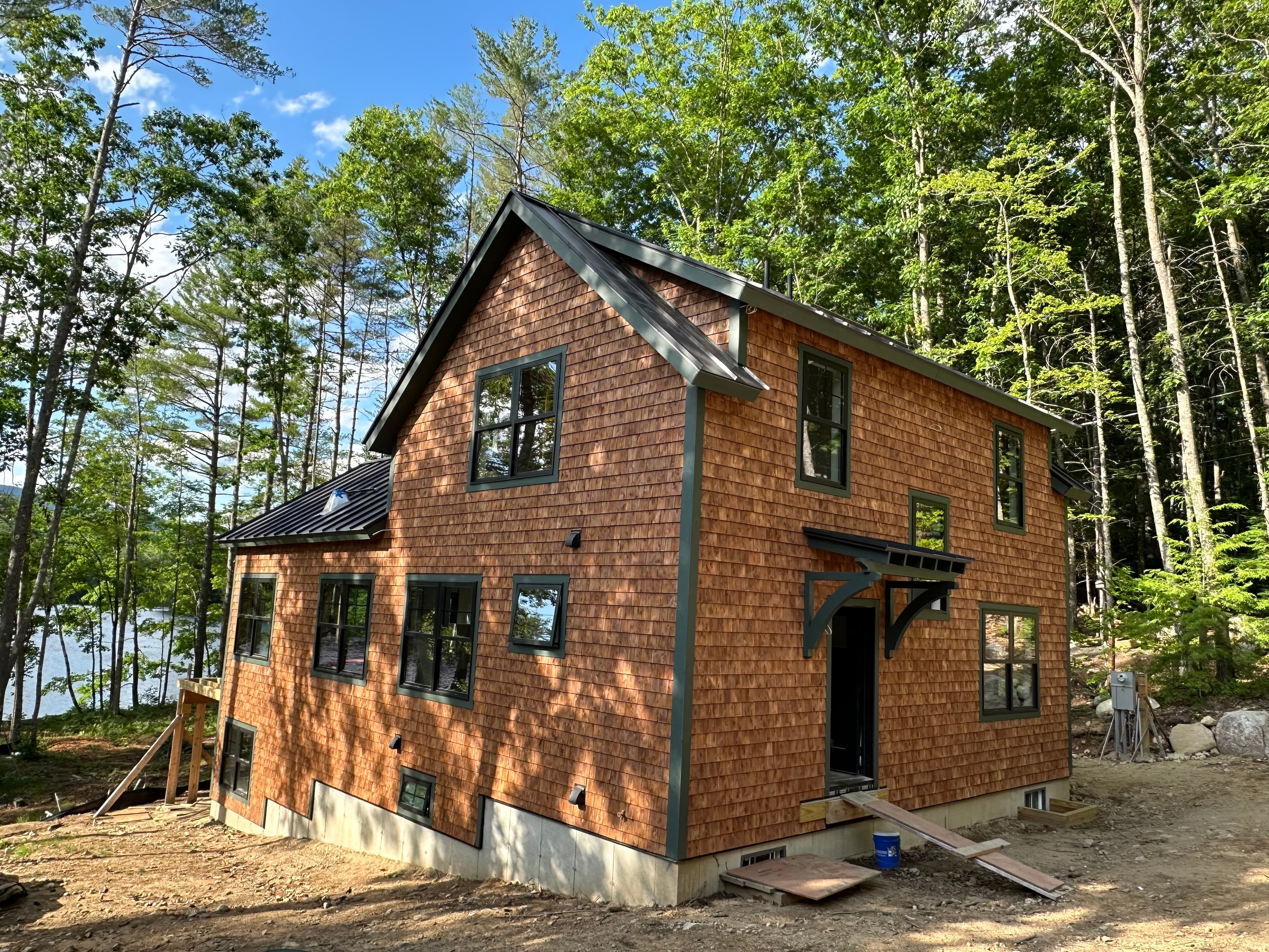 Two-story cedar shingle home with black trim in a sunny, wooded lakeside construction site.
